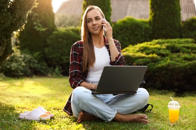 Woman with laptop talking on smartphone on green lawn in park