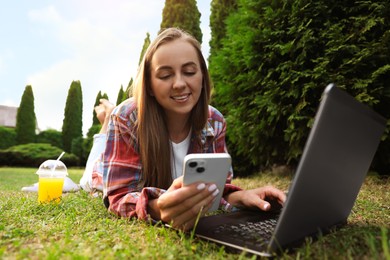 Woman with smartphone using laptop on green lawn in park