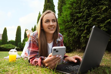 Woman with smartphone using laptop on green lawn in park