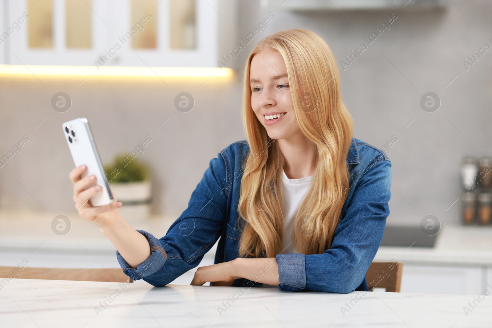 Photo of Smiling woman using smartphone at white table in kitchen