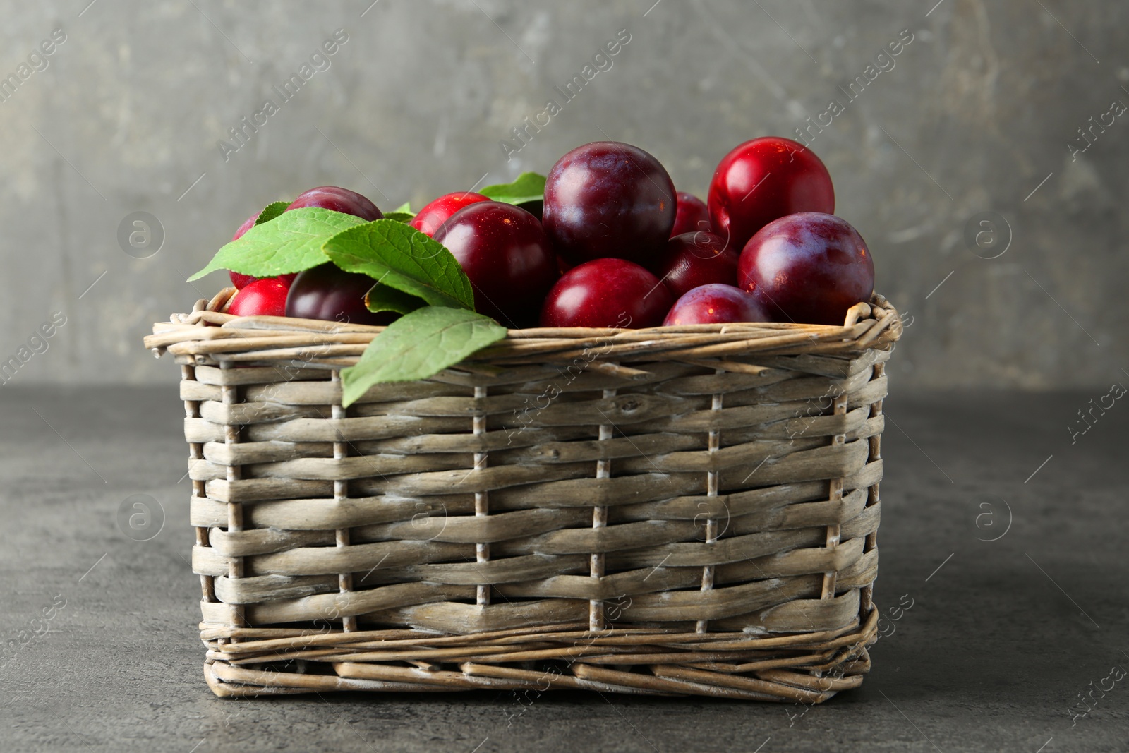 Photo of Tasty ripe plums and leaves in wicker basket on grey textured table