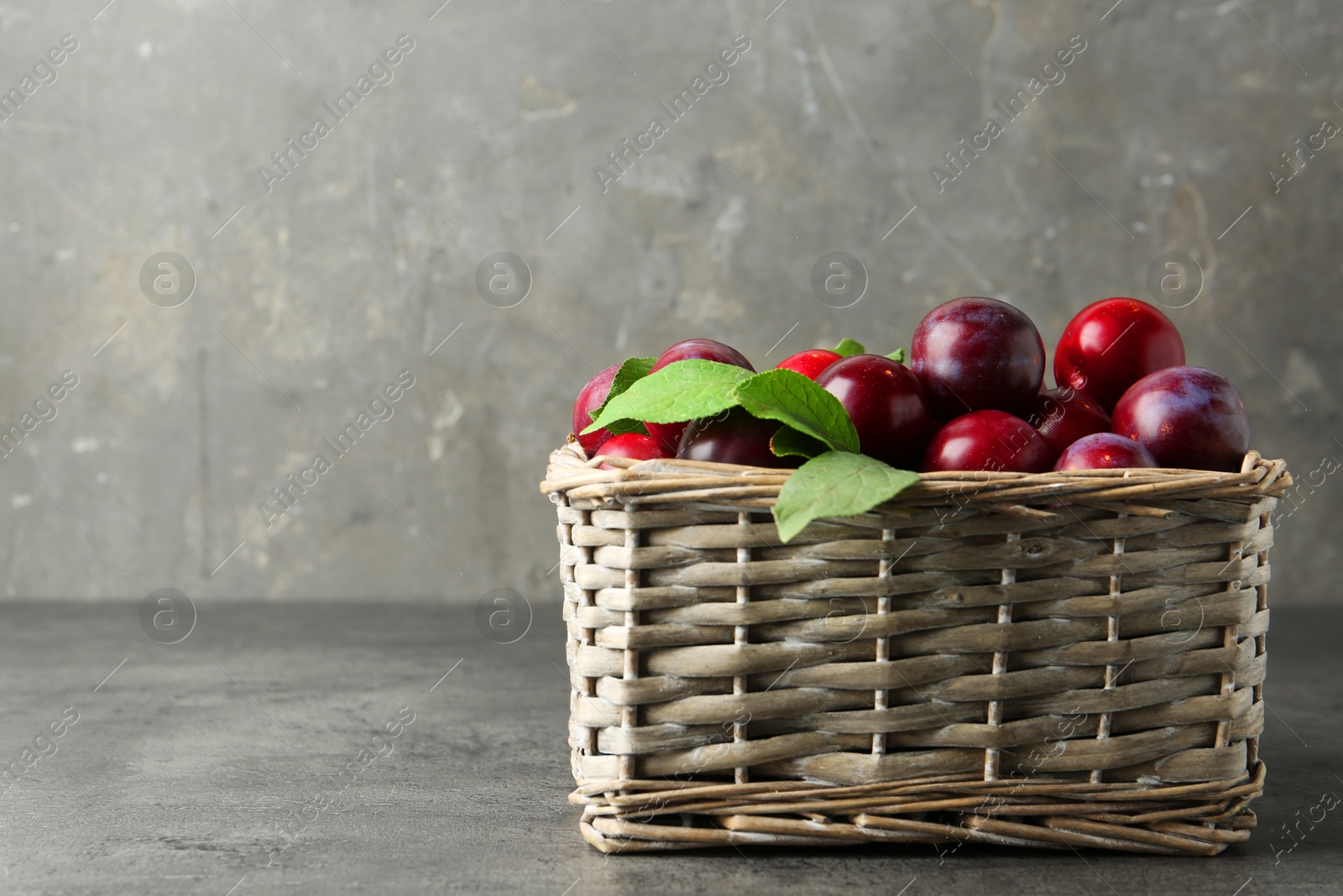 Photo of Tasty ripe plums and leaves in wicker basket on grey textured table, space for text