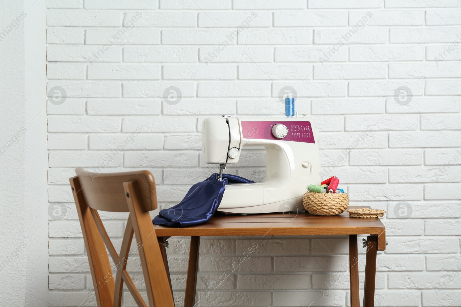 Photo of Sewing machine, blue fabric and spools of threads on wooden table near white brick wall