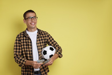 Happy man with money and soccer ball on yellow background, space for text