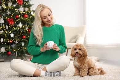 Photo of Woman with cup of drink and cute Maltipoo dog on rug in room decorated for Christmas