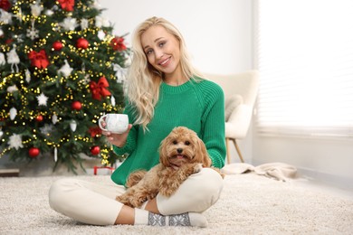 Woman with cup of drink and cute Maltipoo dog on rug in room decorated for Christmas