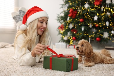 Photo of Woman opening Christmas gift and cute Maltipoo dog on rug indoors