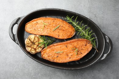 Halves of tasty cooked sweet potato with rosemary and garlic in baking dish on grey textured table, top view