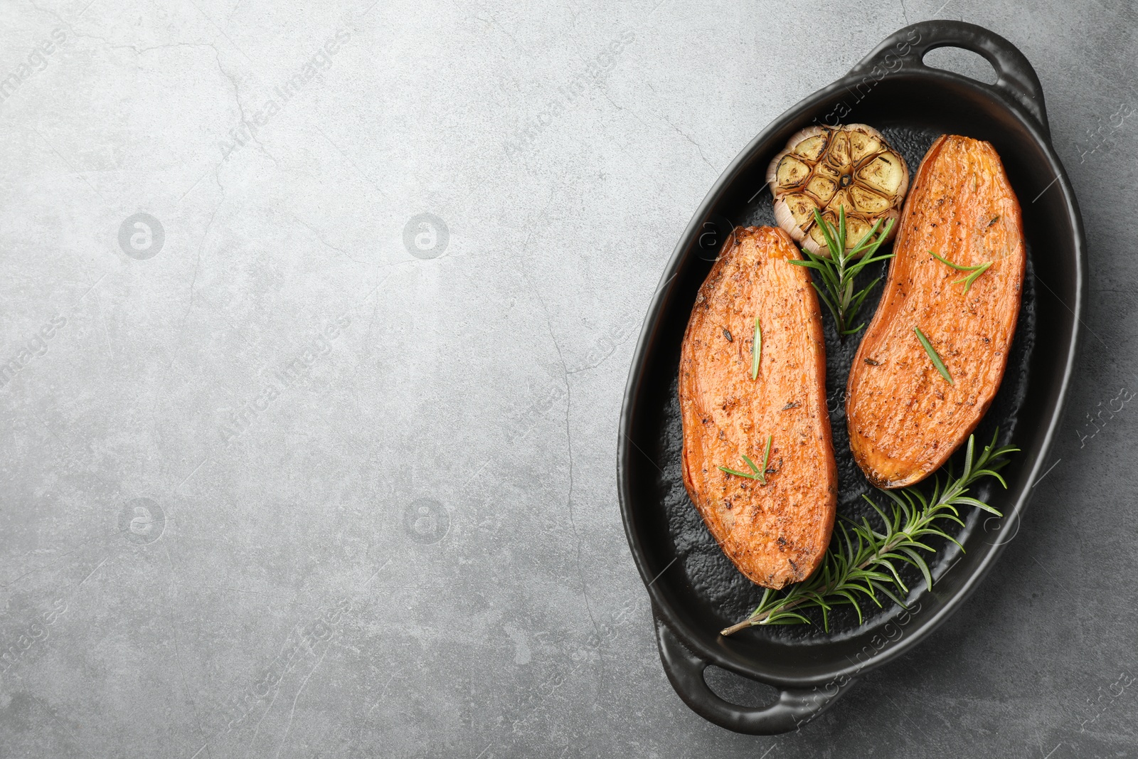 Photo of Halves of tasty cooked sweet potato with rosemary and garlic in baking dish on grey textured table, top view. Space for text