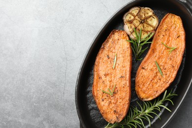 Photo of Halves of tasty cooked sweet potato with rosemary and garlic in baking dish on grey textured table, top view. Space for text