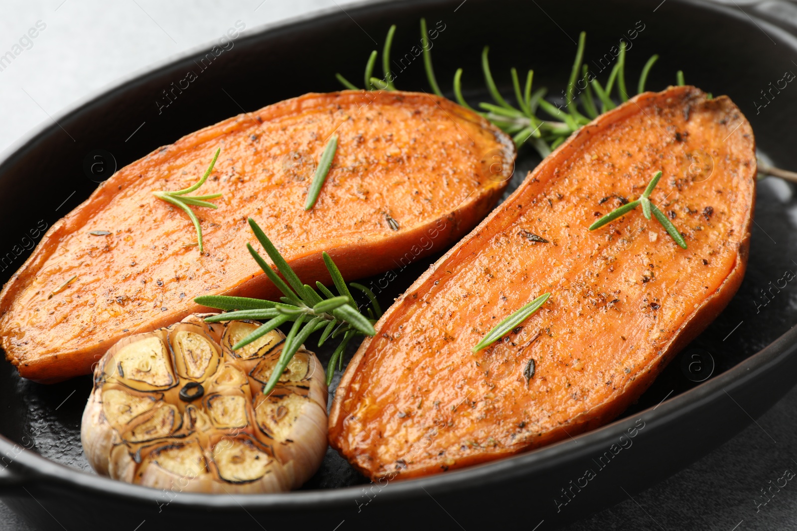 Photo of Halves of tasty cooked sweet potato with rosemary and garlic in baking dish on grey table, closeup