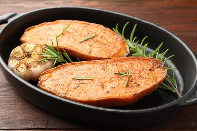 Photo of Halves of tasty cooked sweet potato with rosemary and garlic in baking dish on wooden table, closeup