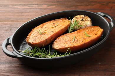 Photo of Halves of tasty cooked sweet potato with rosemary and garlic in baking dish on wooden table, closeup