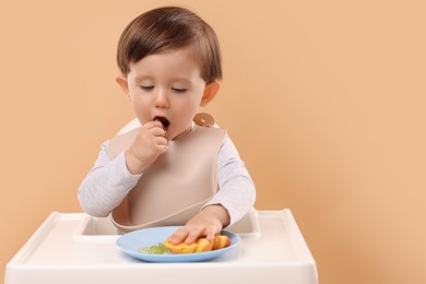 Photo of Healthy baby food. Cute little kid eating fruits in high chair on beige background, space for text