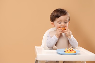 Photo of Healthy baby food. Cute little kid eating fruits in high chair on beige background, space for text