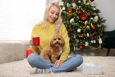 Photo of Woman with cup of drink and cute Maltipoo dog in room decorated for Christmas