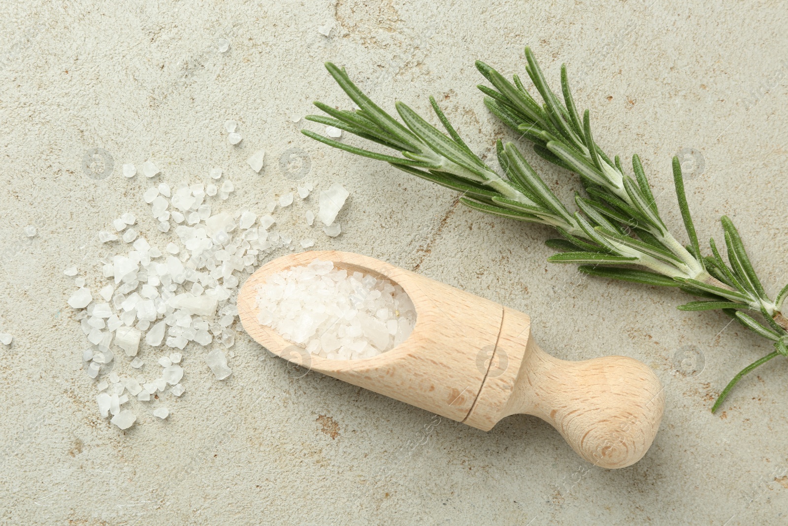 Photo of Sea salt in wooden scoop and rosemary on grey table, top view