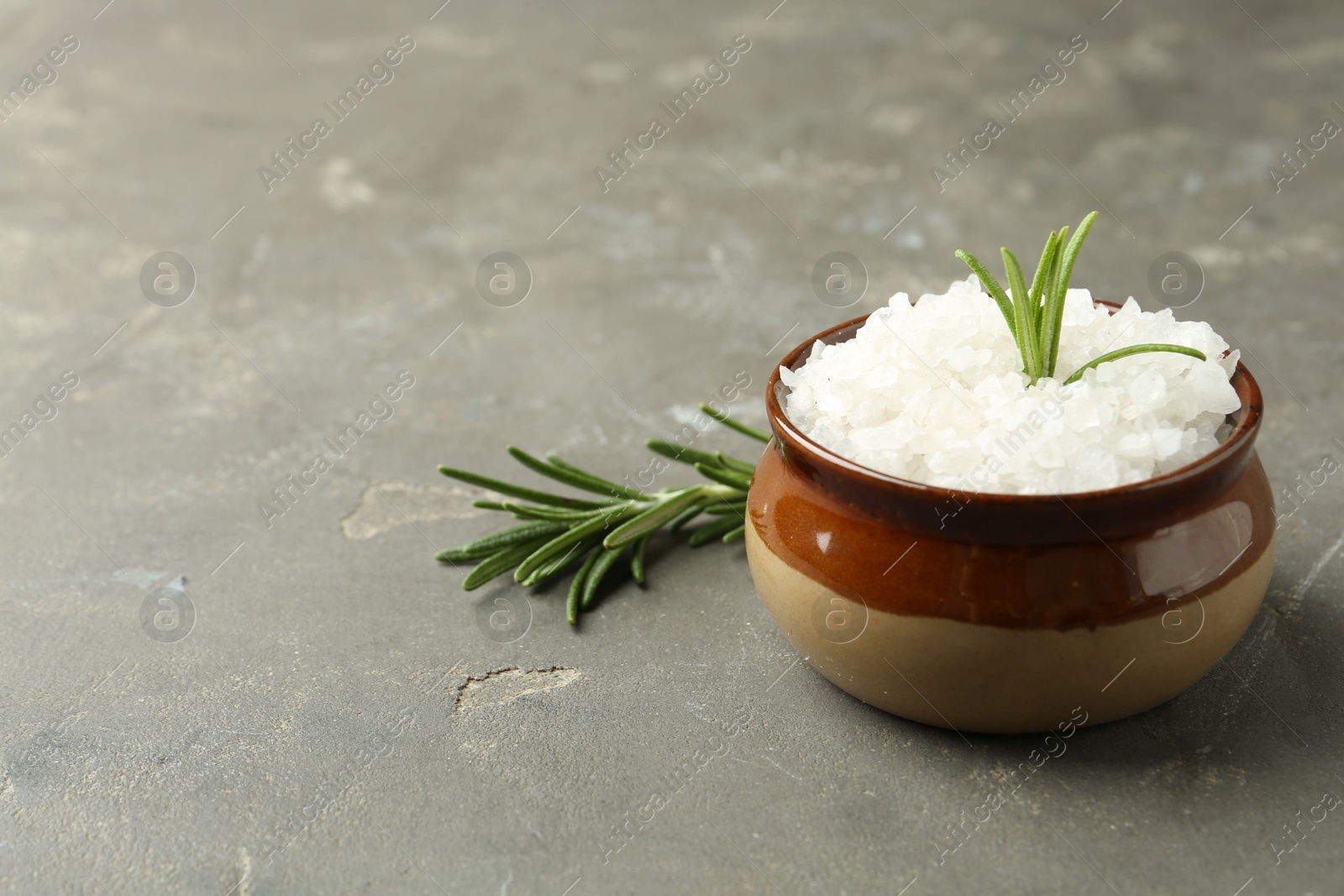 Photo of Sea salt in bowl and rosemary on grey table, closeup. Space for text