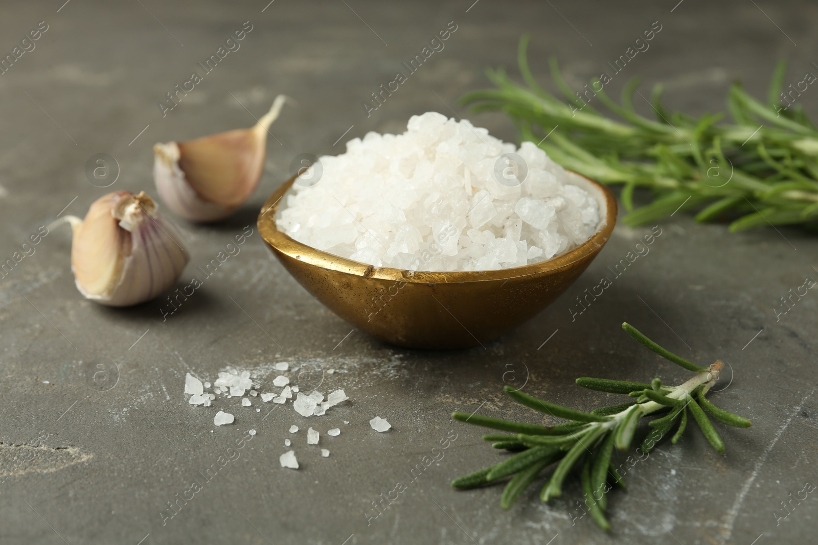 Photo of Sea salt in bowl, rosemary and garlic on grey table, closeup