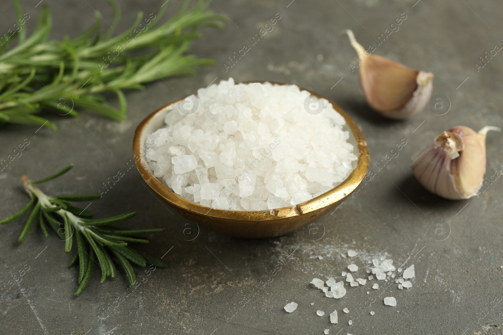 Photo of Sea salt in bowl, rosemary and garlic on grey table, closeup