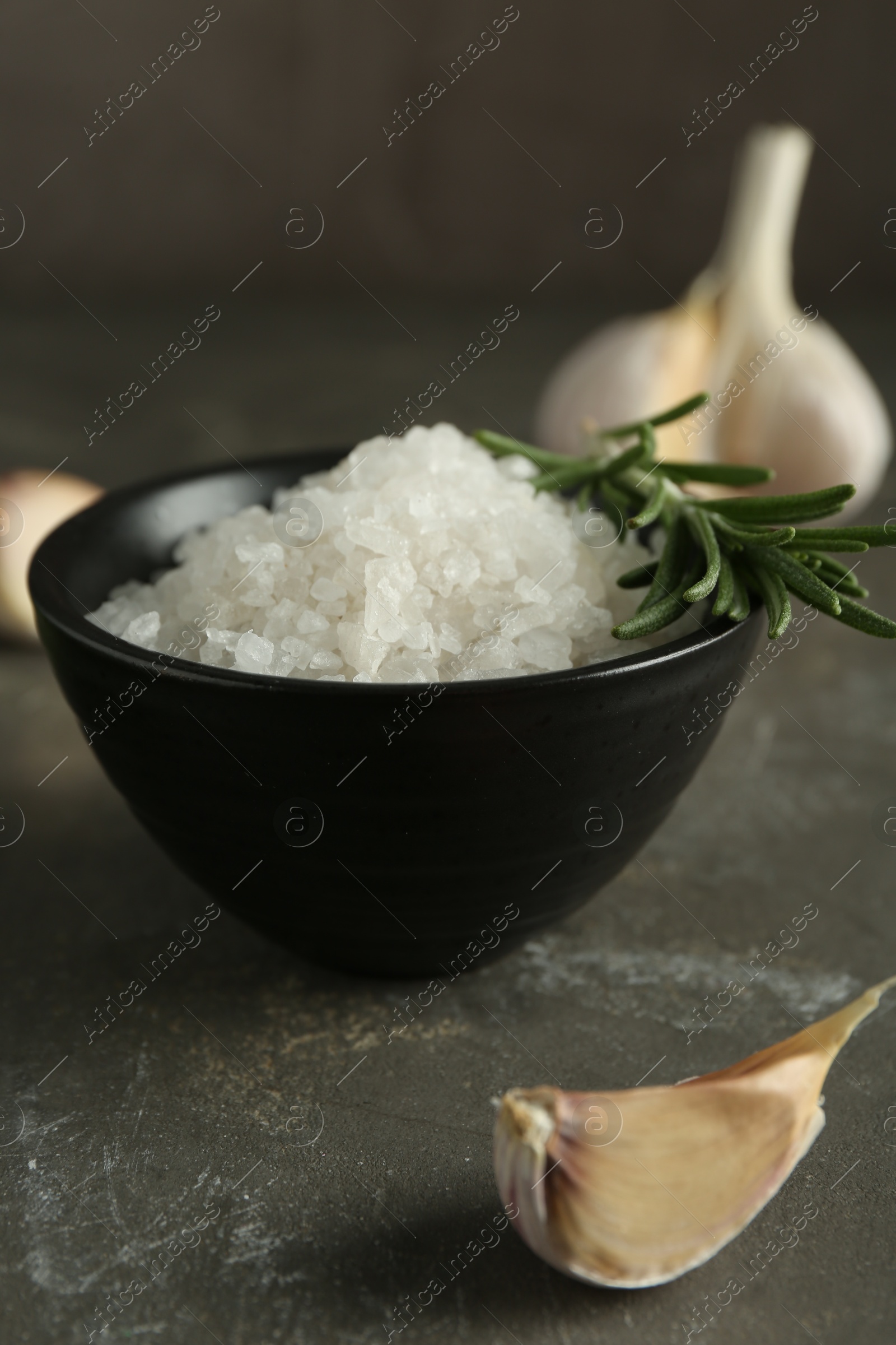 Photo of Sea salt in bowl, rosemary and garlic on grey table, closeup