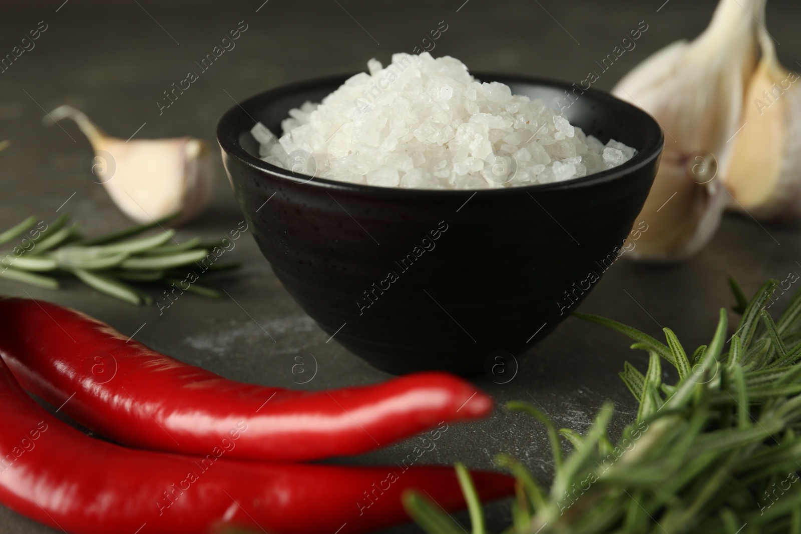 Photo of Sea salt in bowl, rosemary, chili peppers and garlic on grey table, closeup