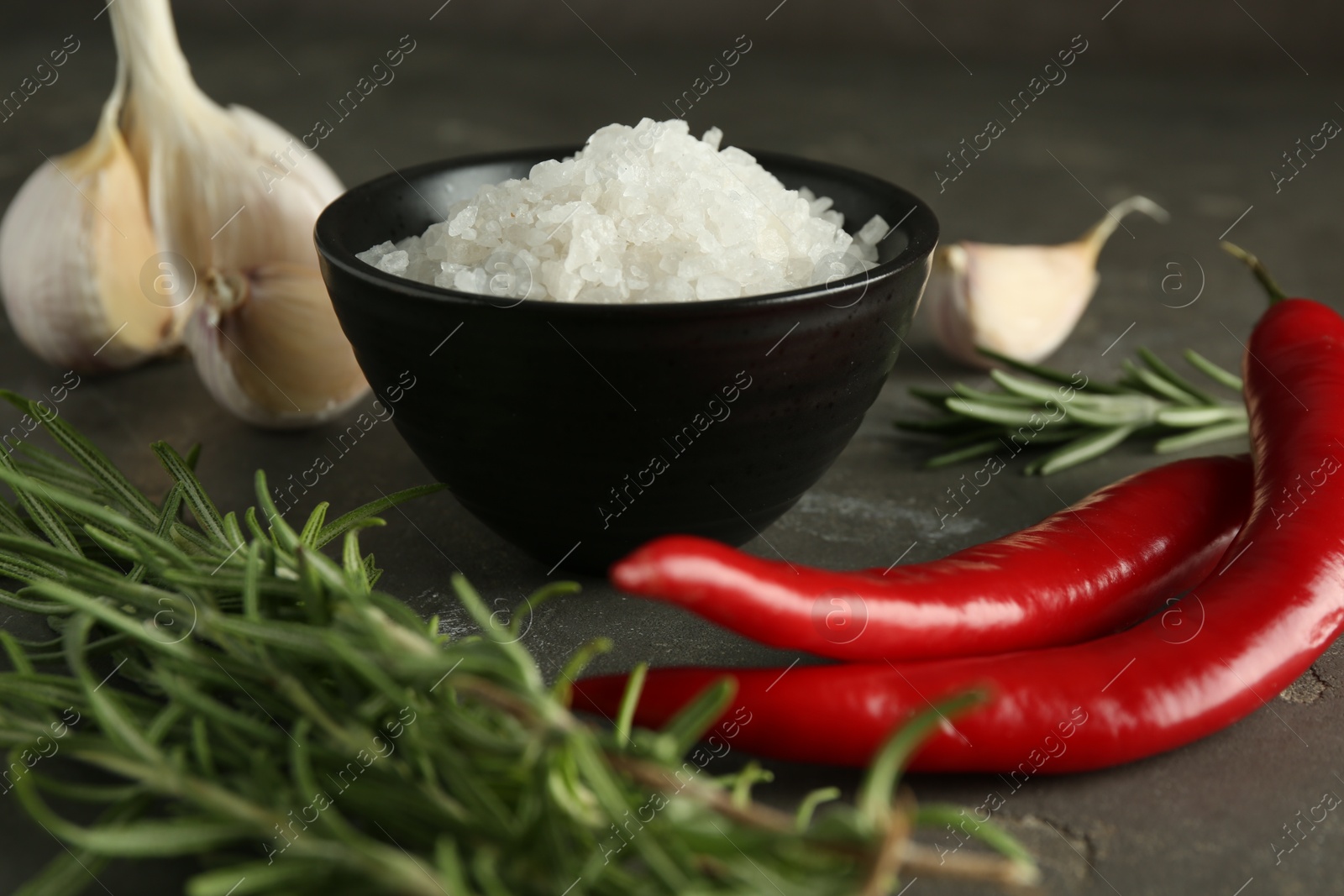 Photo of Sea salt in bowl, rosemary, chili peppers and garlic on grey table, closeup