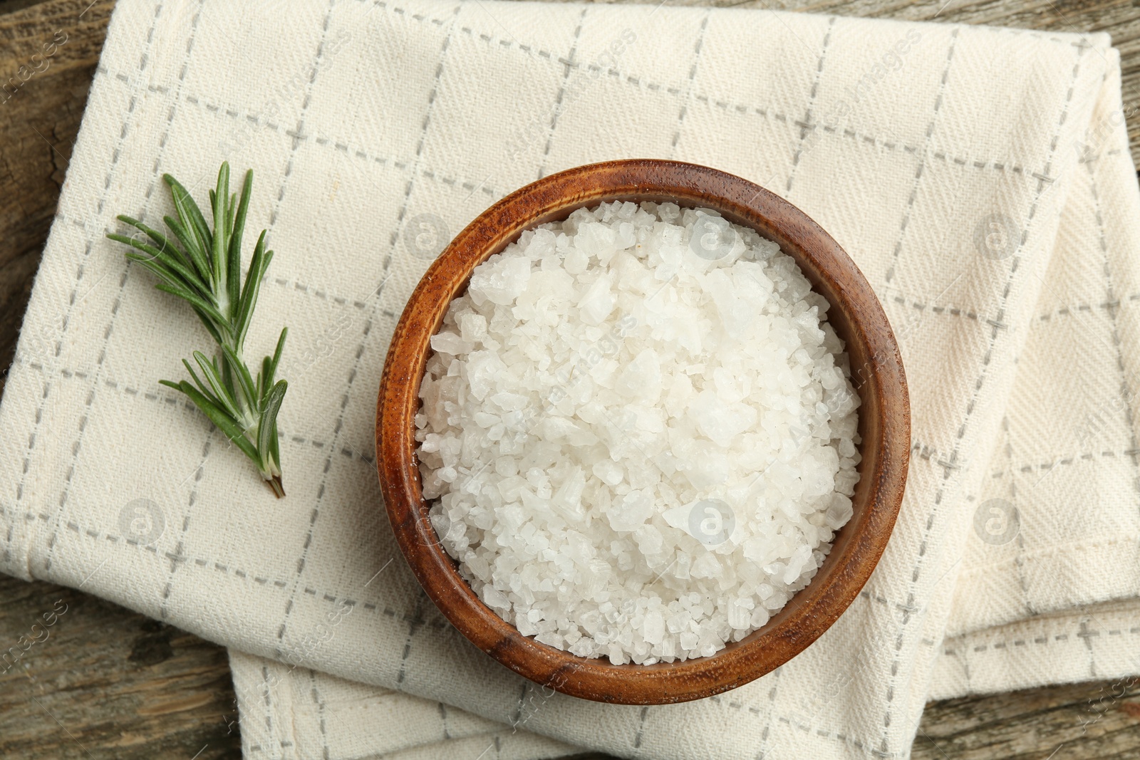 Photo of Sea salt in bowl and rosemary on wooden table, top view