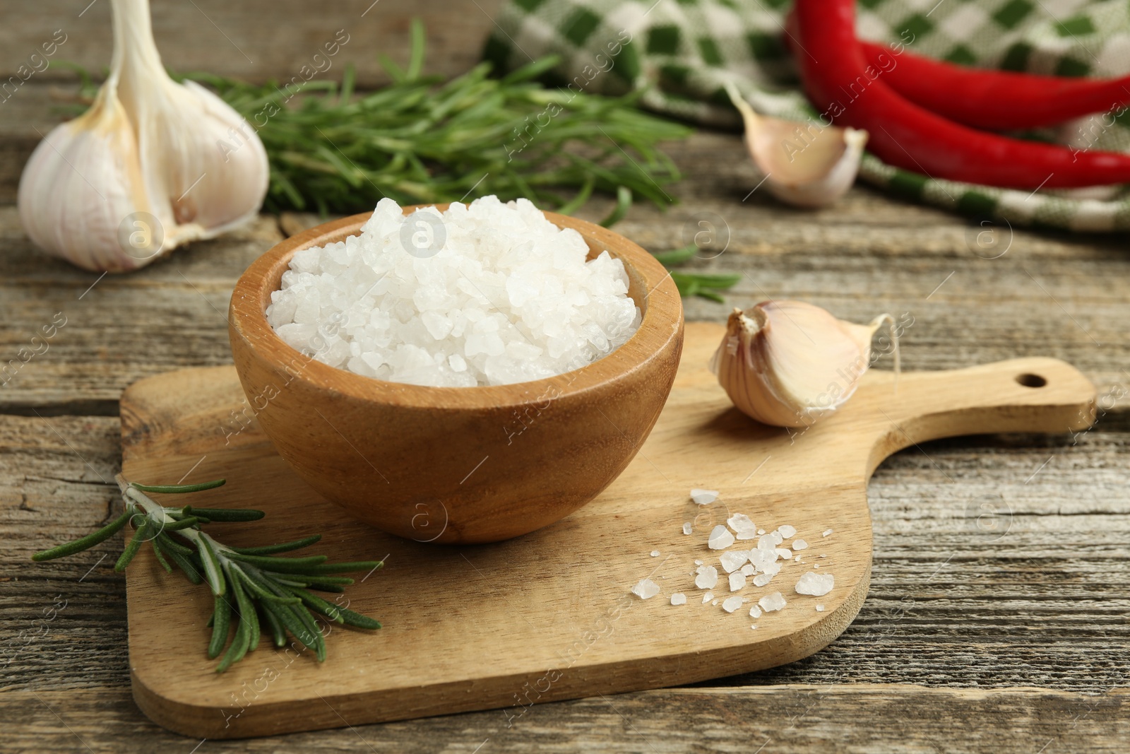 Photo of Sea salt in bowl, rosemary and garlic on wooden table, closeup