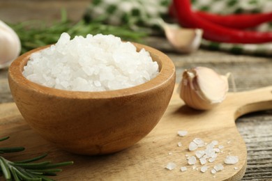 Photo of Sea salt in bowl, rosemary and garlic on wooden table, closeup