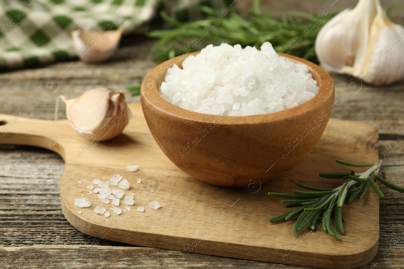 Photo of Sea salt in bowl, rosemary and garlic on wooden table, closeup