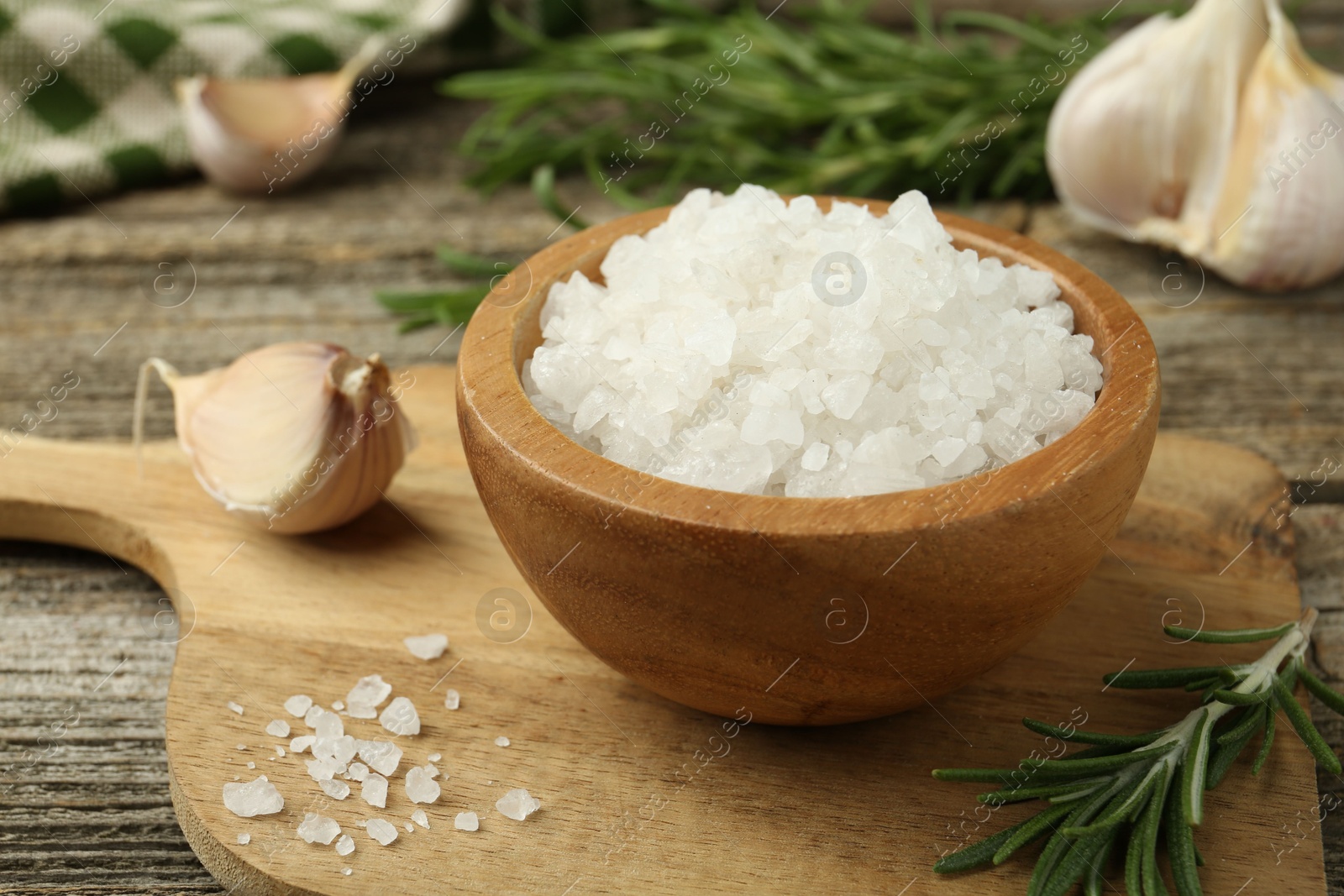 Photo of Sea salt in bowl, rosemary and garlic on wooden table, closeup