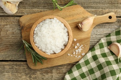 Photo of Sea salt in bowl, rosemary and garlic on wooden table, top view