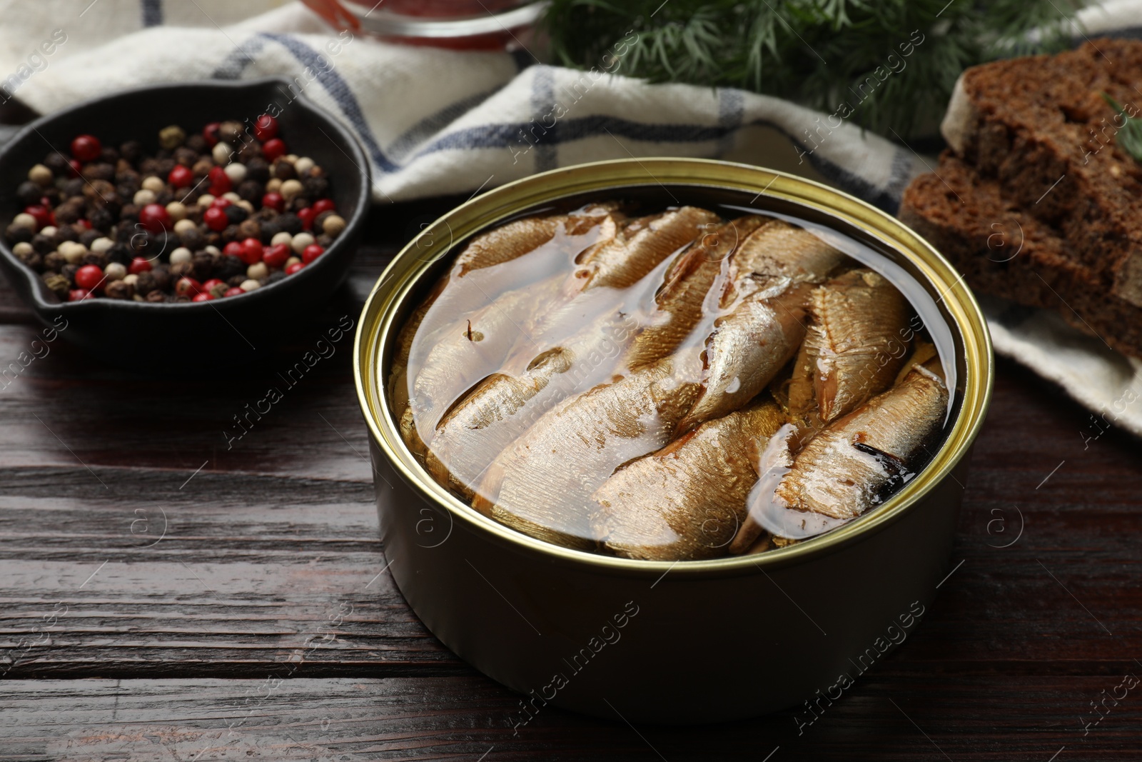 Photo of Tasty sprats in tin can, peppercorns, bread and dill on wooden table, closeup