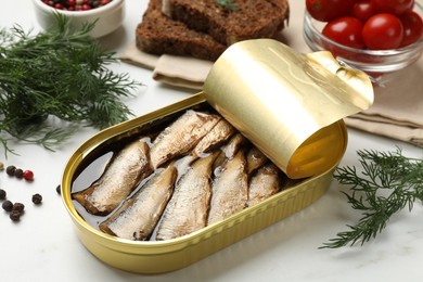 Tasty sprats in tin can, dill, peppercorns, bread and tomatoes on white table, closeup