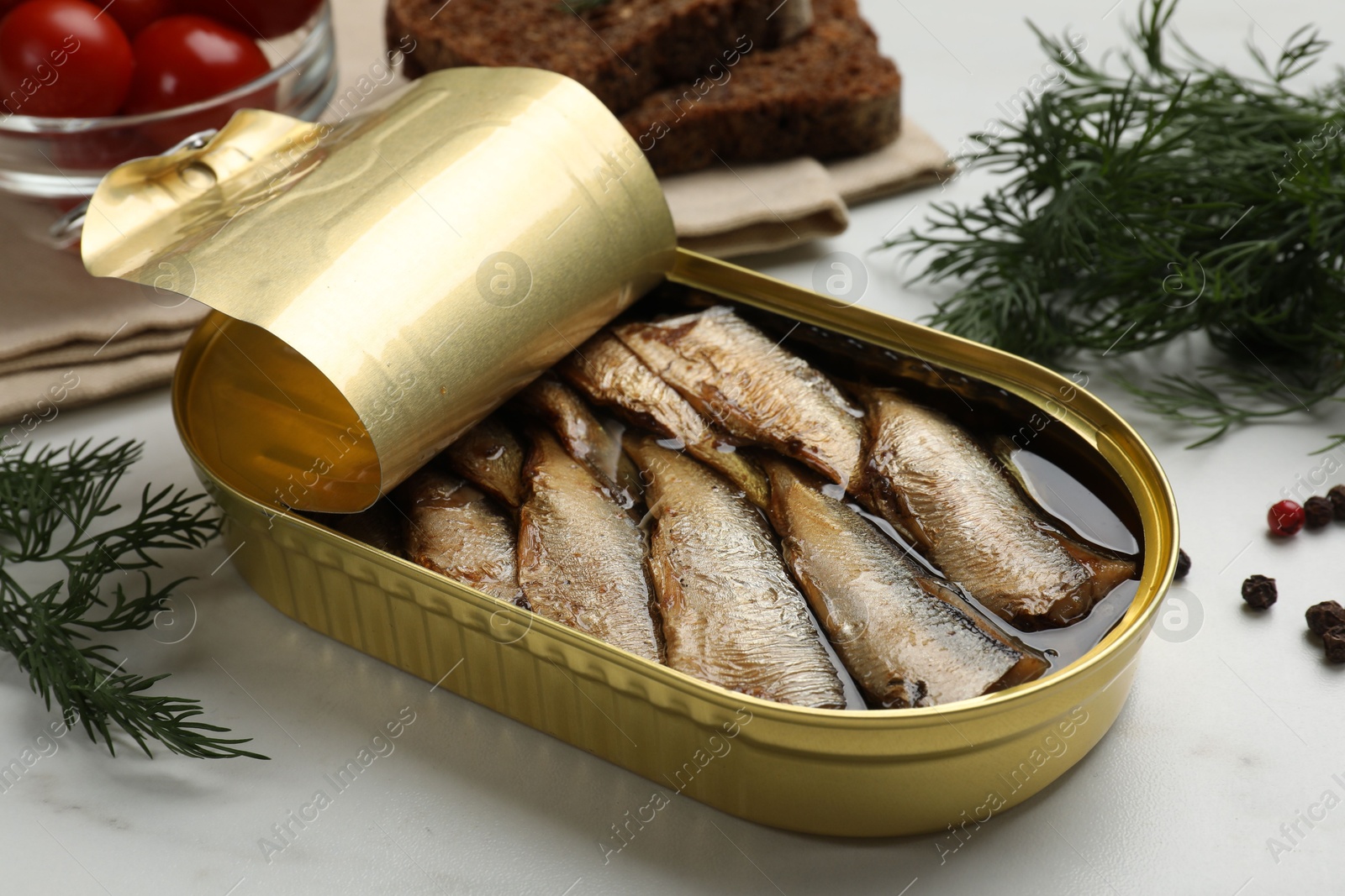 Photo of Tasty sprats in tin can, dill, peppercorns, bread and tomatoes on white table, closeup