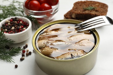 Photo of Tasty sprats in tin can, dill, peppercorns, bread and tomatoes on white table, closeup