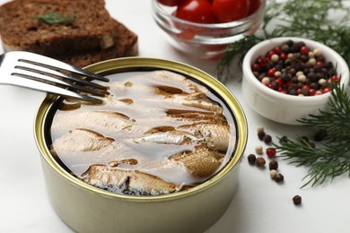 Tasty sprats in tin can, dill, peppercorns, bread and tomatoes on white table, closeup