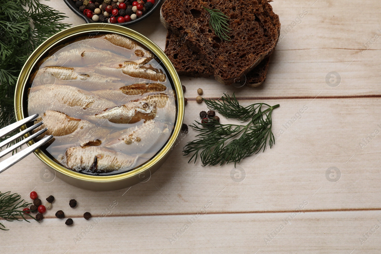 Photo of Tasty sprats in tin can, dill, bread and peppercorns on wooden table, top view