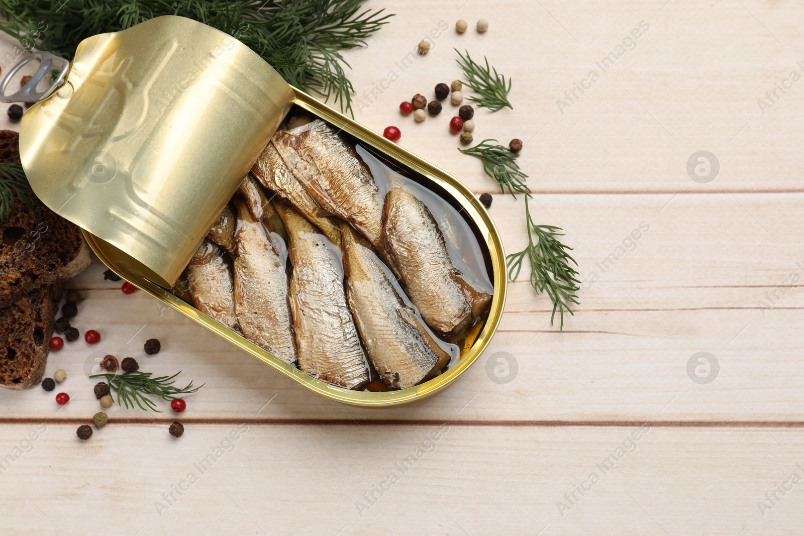 Photo of Tasty sprats in tin can, dill, bread and peppercorns on wooden table, top view