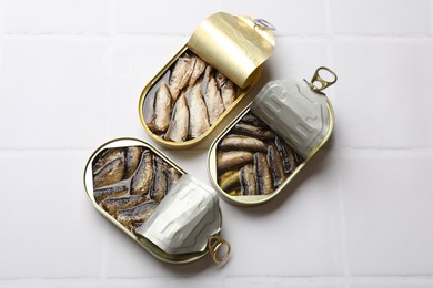 Tasty sprats in tin cans on white tiled table, flat lay