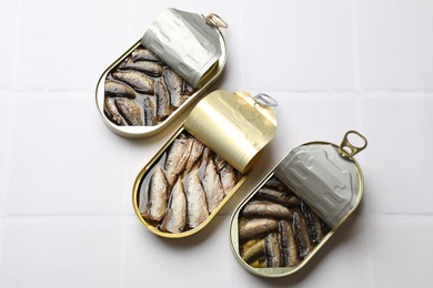 Tasty sprats in tin cans on white tiled table, flat lay