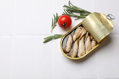 Photo of Tasty sprats in tin can with fresh rosemary and tomato on white tiled table, flat lay. Space for text