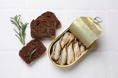 Photo of Tasty sprats in tin can served on white tiled table, flat lay