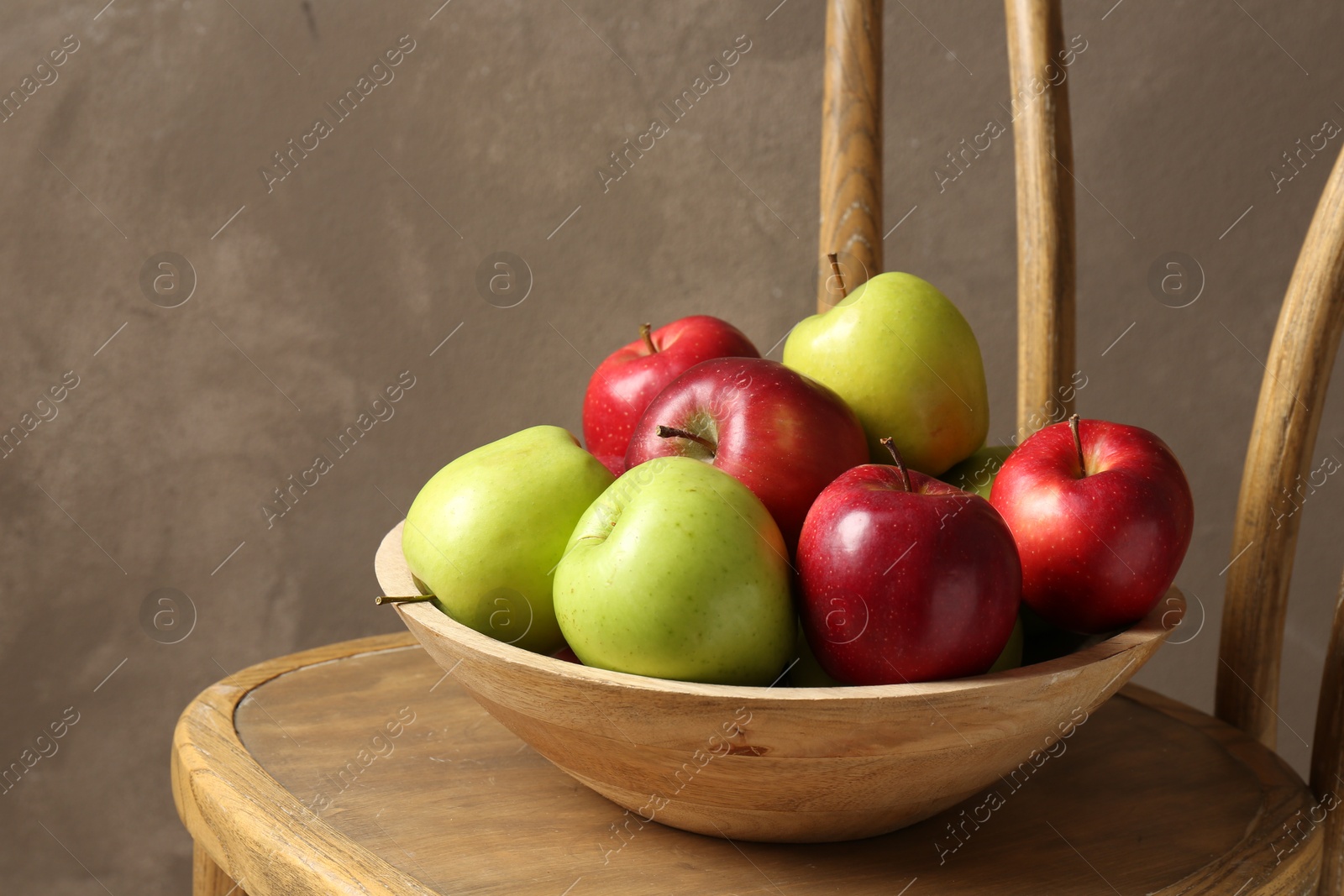 Ripe red and green apples in bowl on wooden chair near grey wall Photo of Ripe red and green apples in bowl on wooden chair near grey wall