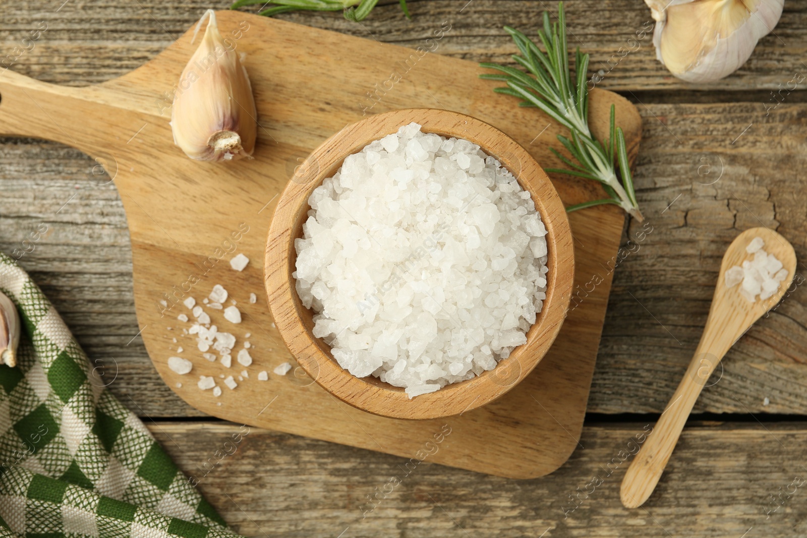 Photo of Sea salt in bowl, rosemary and garlic on wooden table, top view