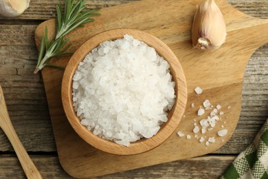 Photo of Sea salt in bowl, rosemary and garlic on wooden table, top view