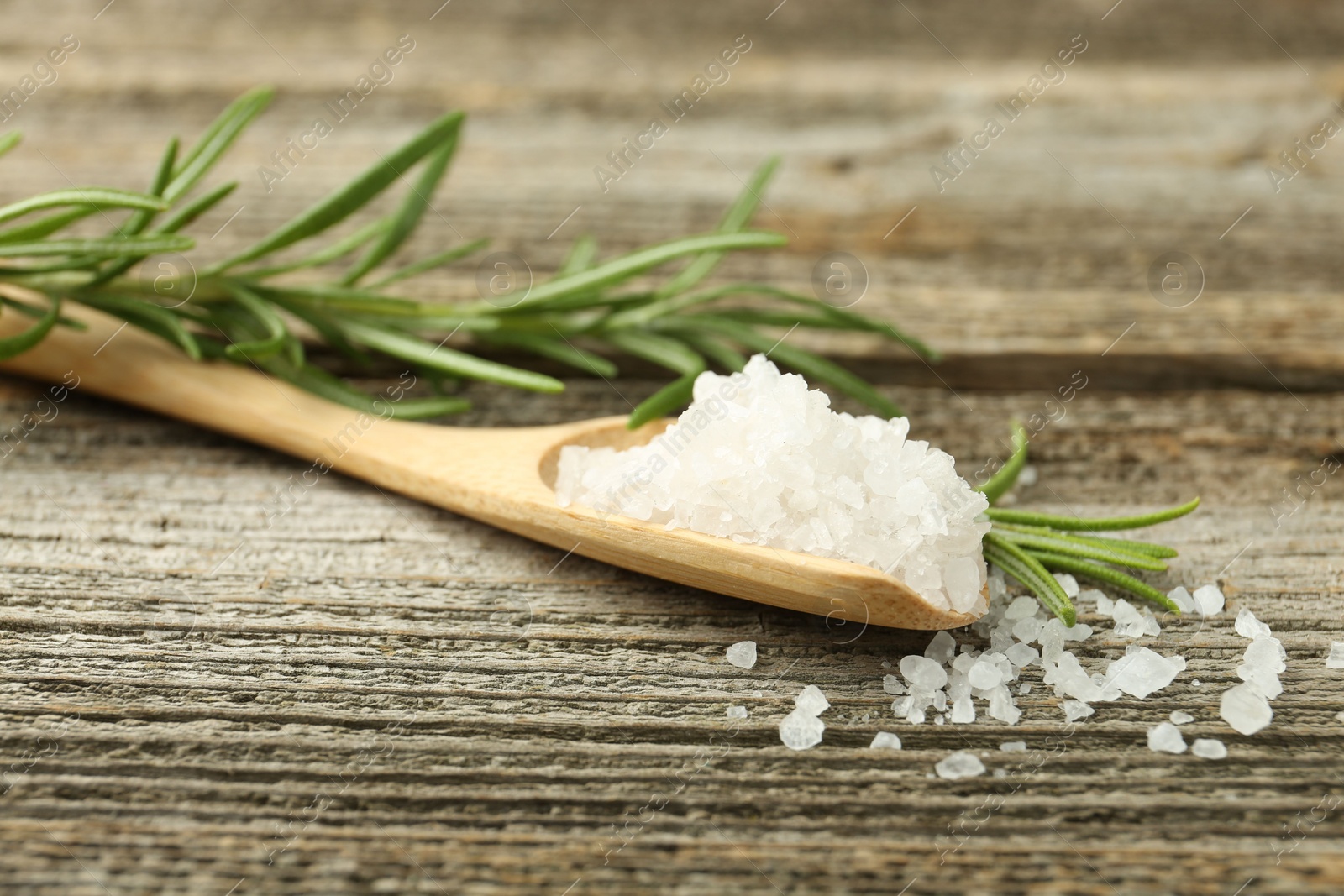 Photo of Sea salt in spoon and rosemary on wooden table, closeup