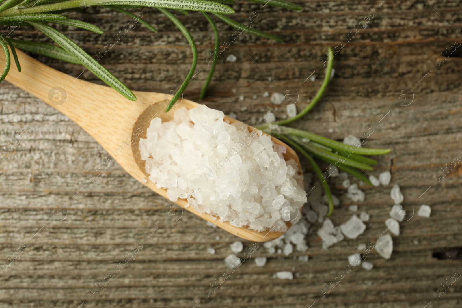 Photo of Sea salt in spoon and rosemary on wooden table, top view