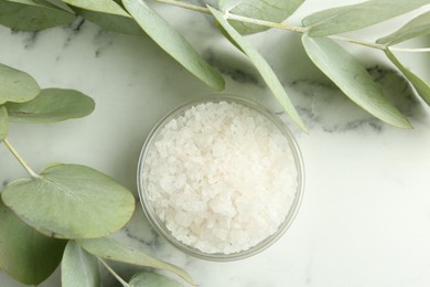 Photo of Sea salt in glass bowl and eucalyptus leaves on white marble table, top view